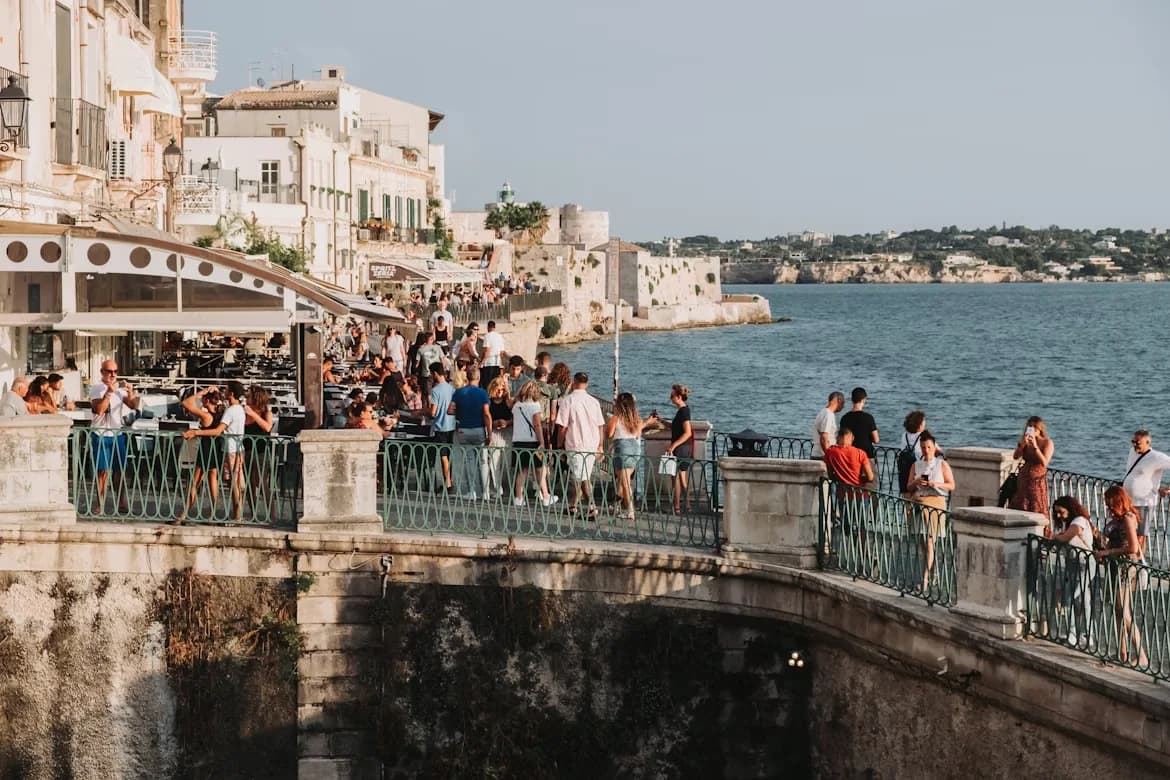 People dine at outdoor cafes on a seaside promenade, enjoying the view of the tranquil sea and the historic buildings of the city.