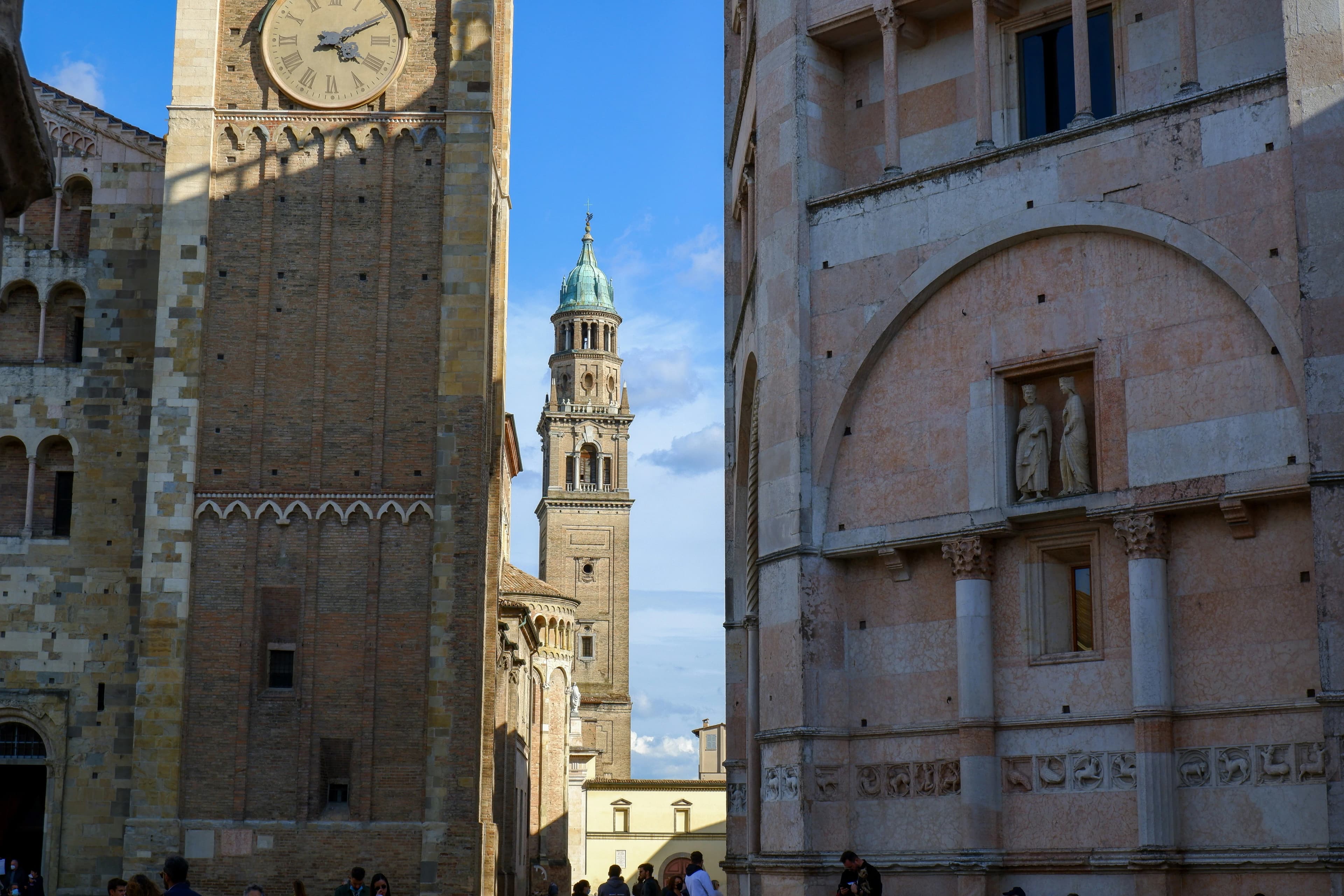 The magnificent bell tower of the Parma Cathedral is viewed from a narrow street, framed by the historic buildings of the city.