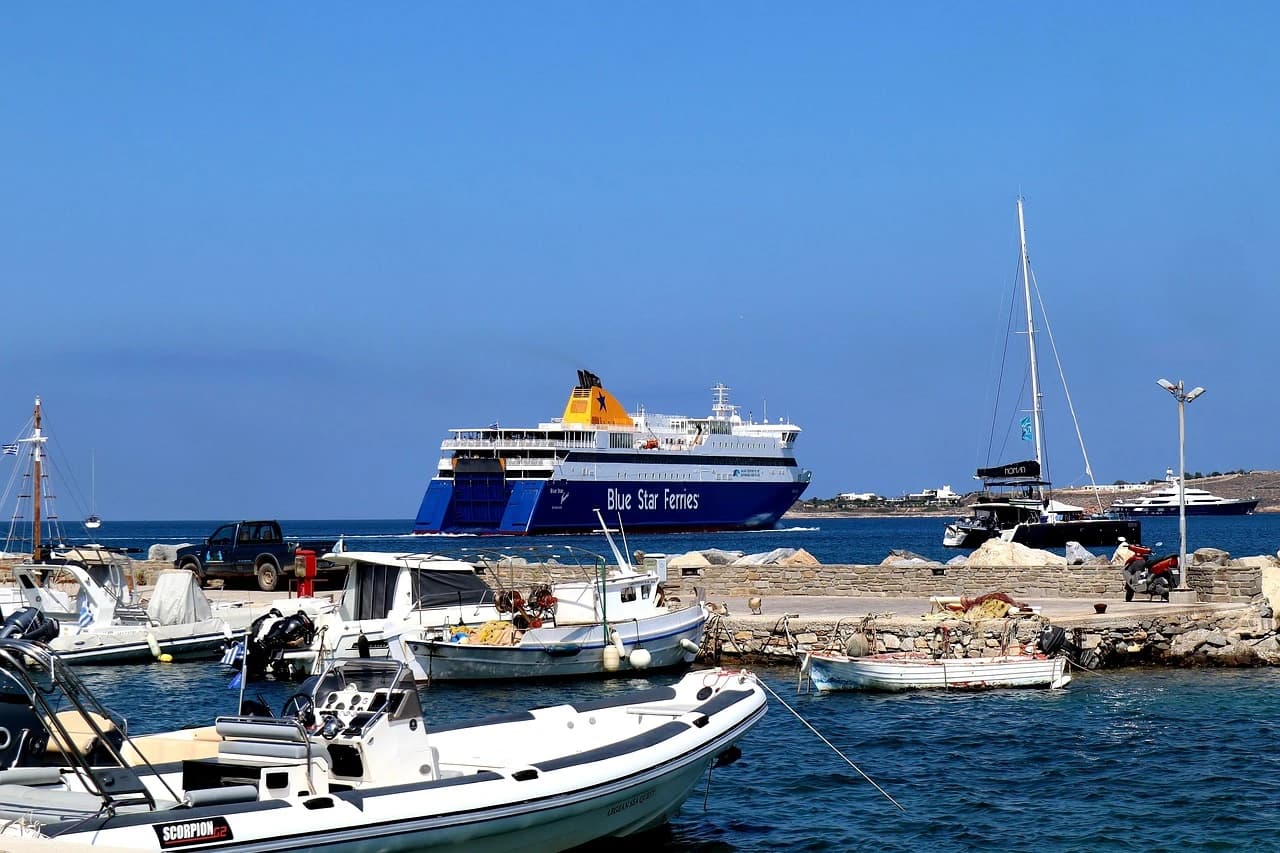 A large, blue ferry with "Blue Star Ferries" written on the side pulls into the harbor, with sailboats and fishing boats moored nearby.