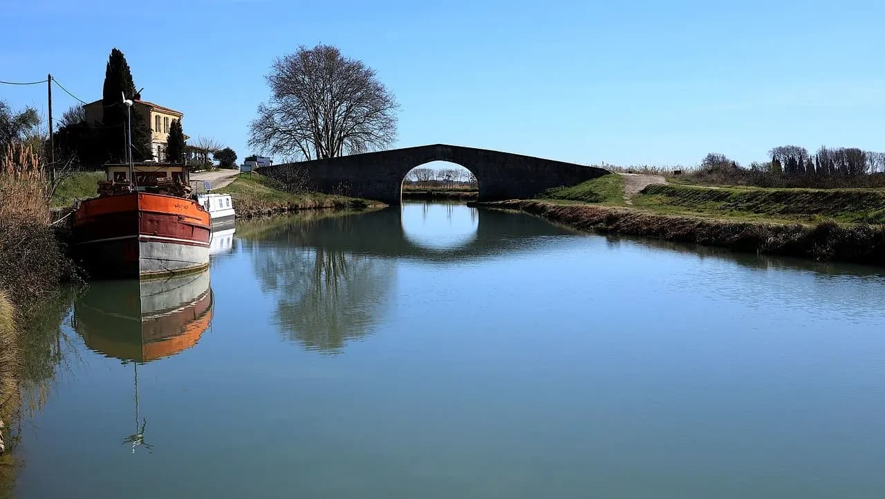 A small fishing boat is moored on a calm river with a historic stone bridge in the background, its arches reflected in the water.