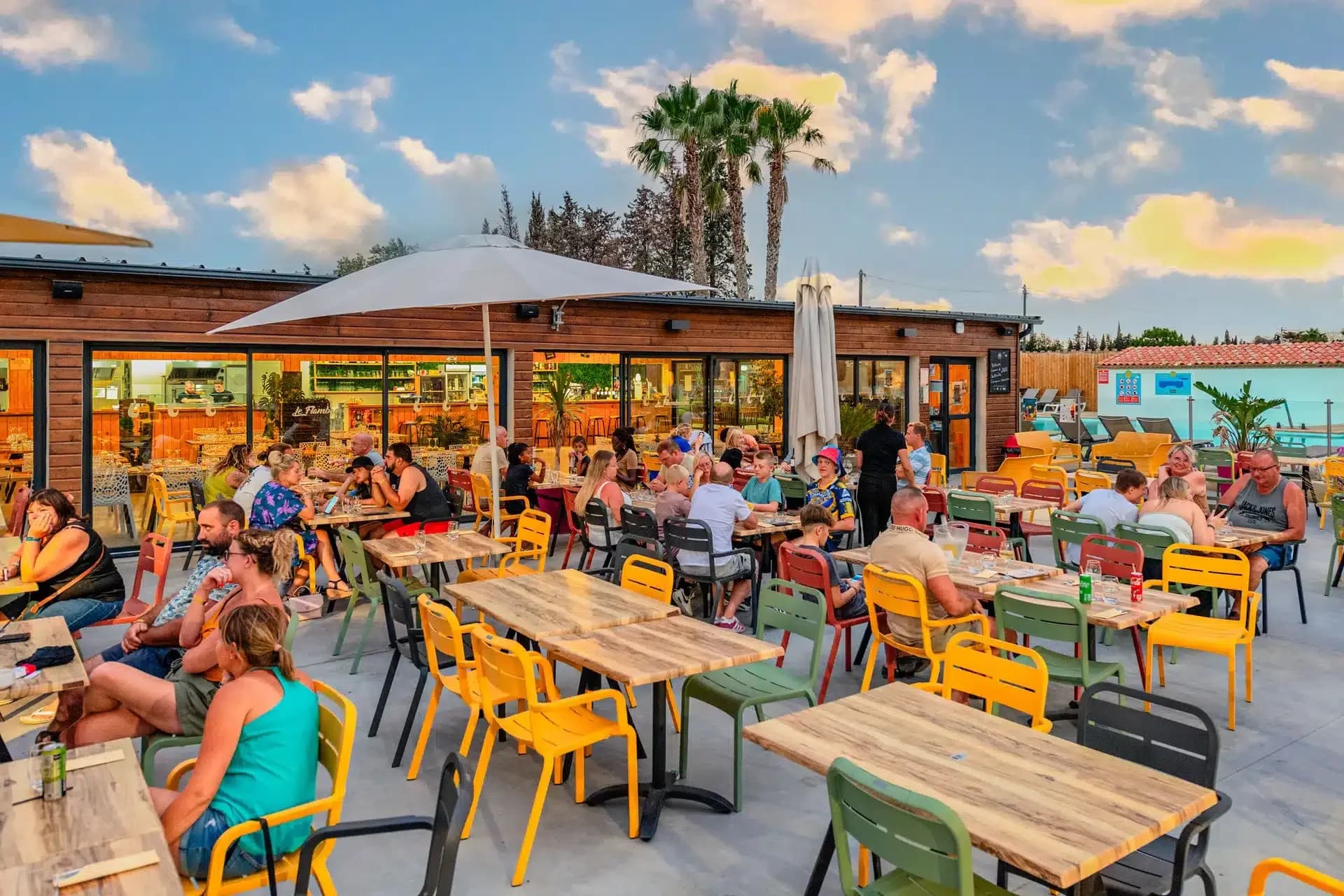 A vibrant outdoor restaurant is bustling with people enjoying a meal at sunset, with palm trees and a colorful sky in the background.