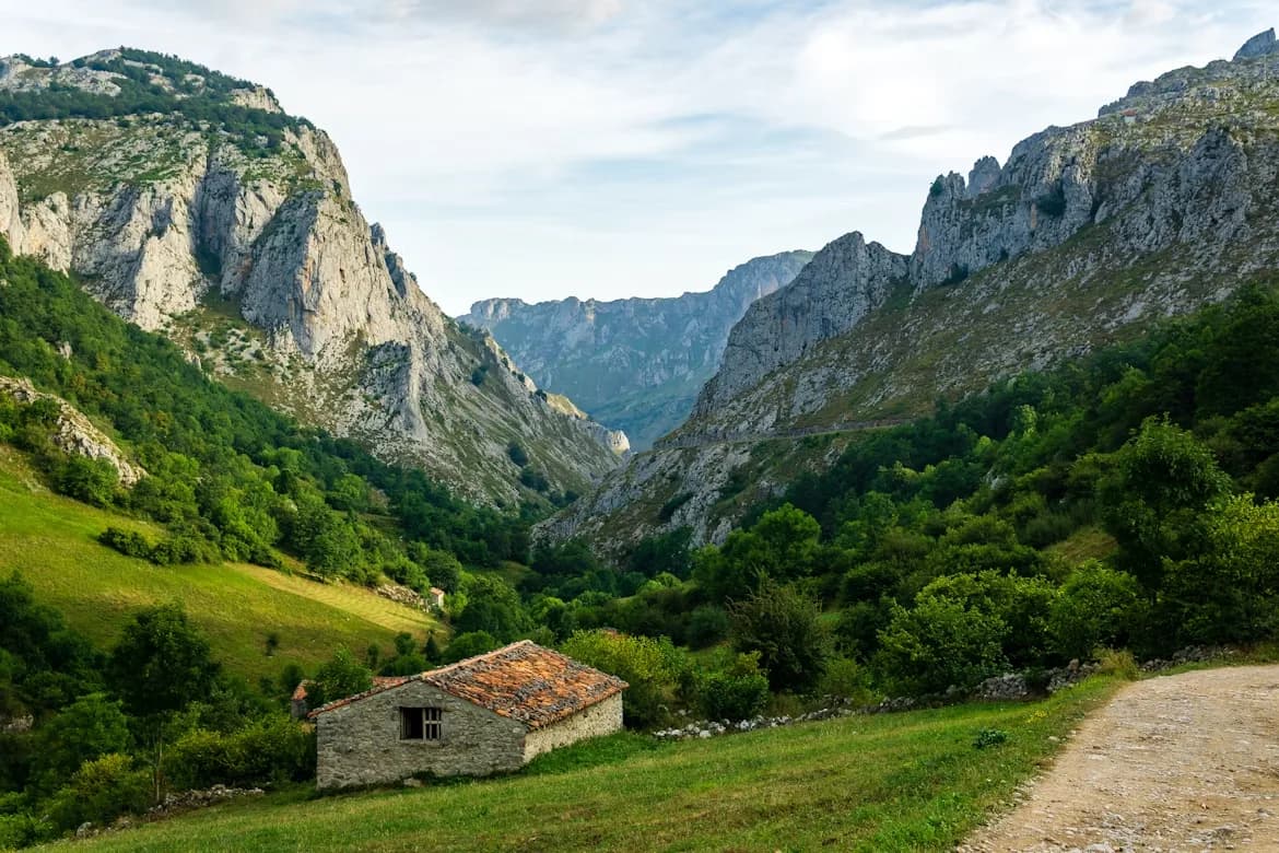 A small, rustic stone house sits in a lush green valley, with towering, rocky cliffs on either side.