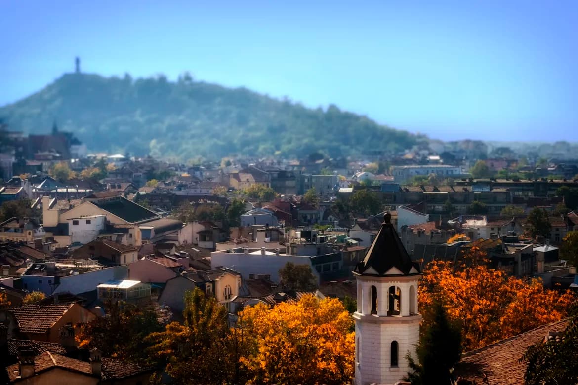 A small bell tower with a pointed roof rises above the rooftops of Plovdiv's historic Old Town, with a large, forested hill in the background.