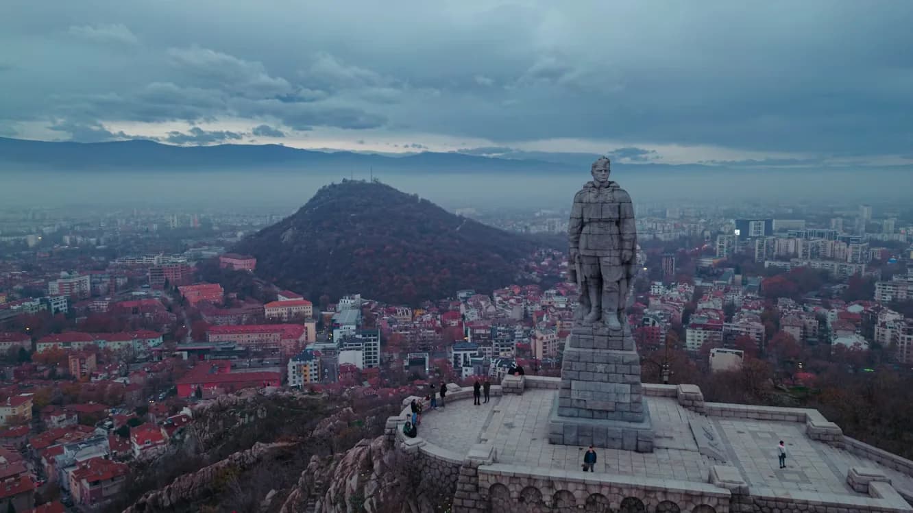 A large, grey stone statue of a soldier stands on a hill, overlooking the city of Plovdiv, with a cloudy sky in the background.