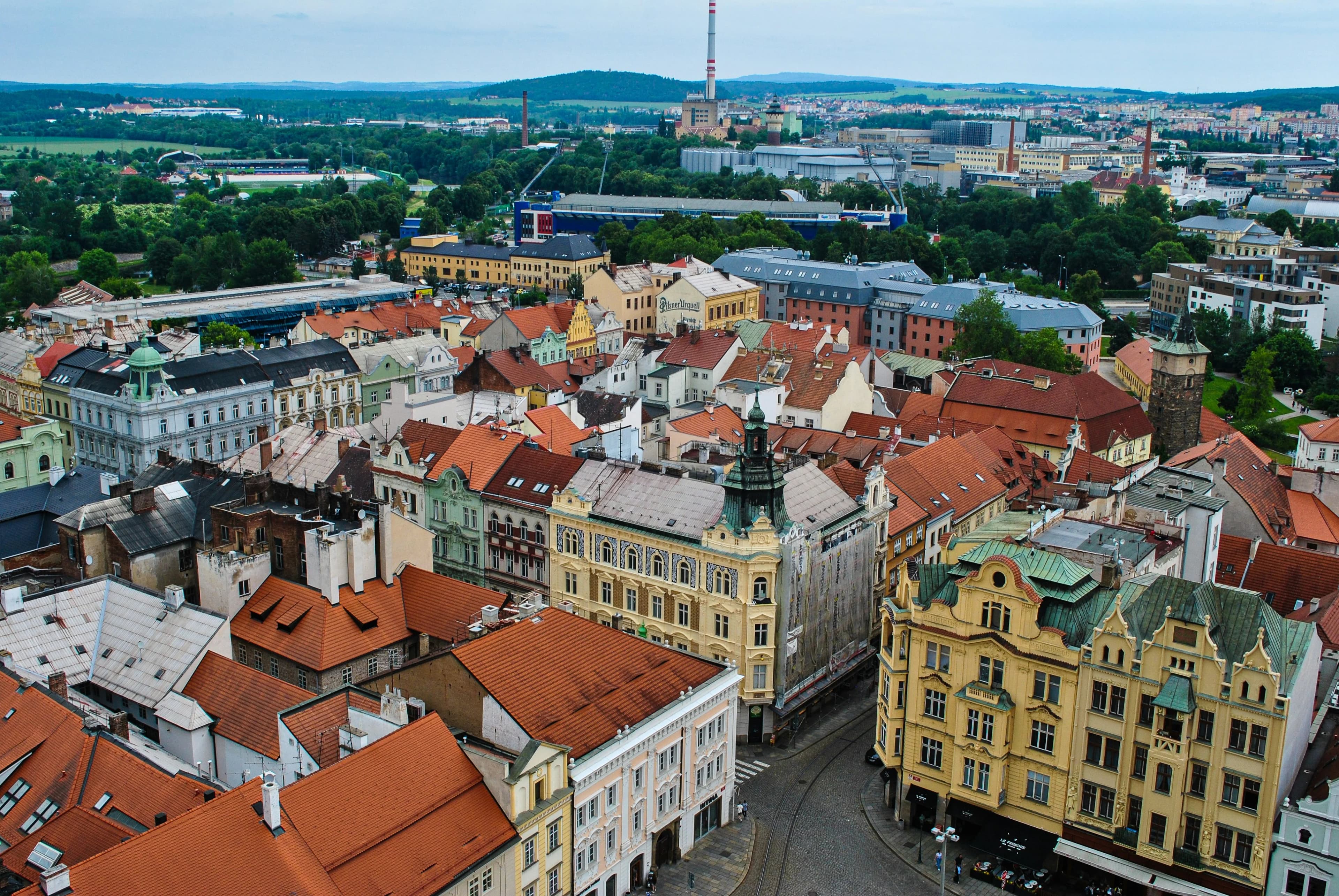 An aerial view captures the city of Plzeň, with its traditional red-tiled roofs and historic buildings, with a large industrial complex in the background.