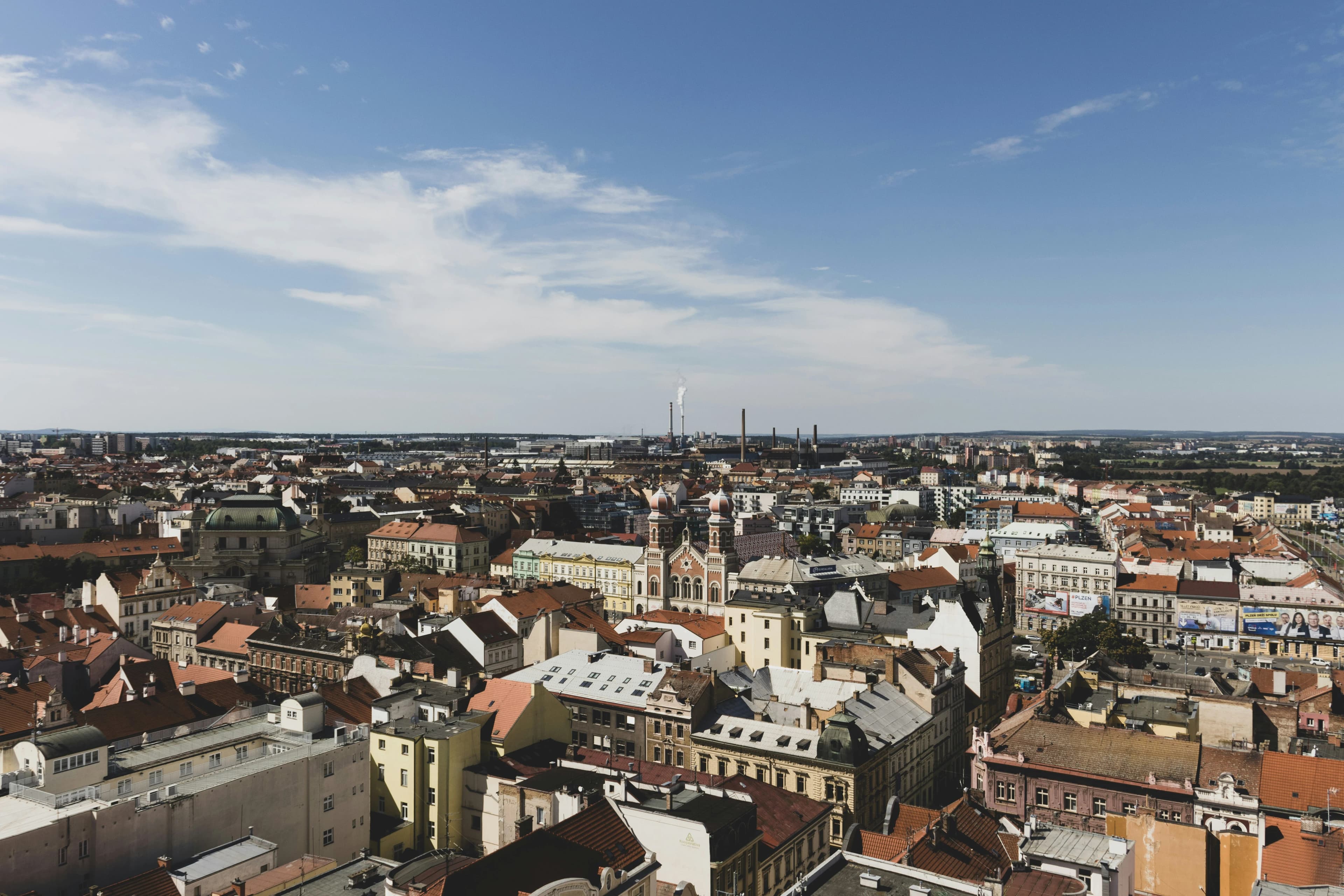 A panoramic view of the Plzeň city skyline shows a dense cluster of historic buildings and rooftops, with industrial chimneys in the distance.