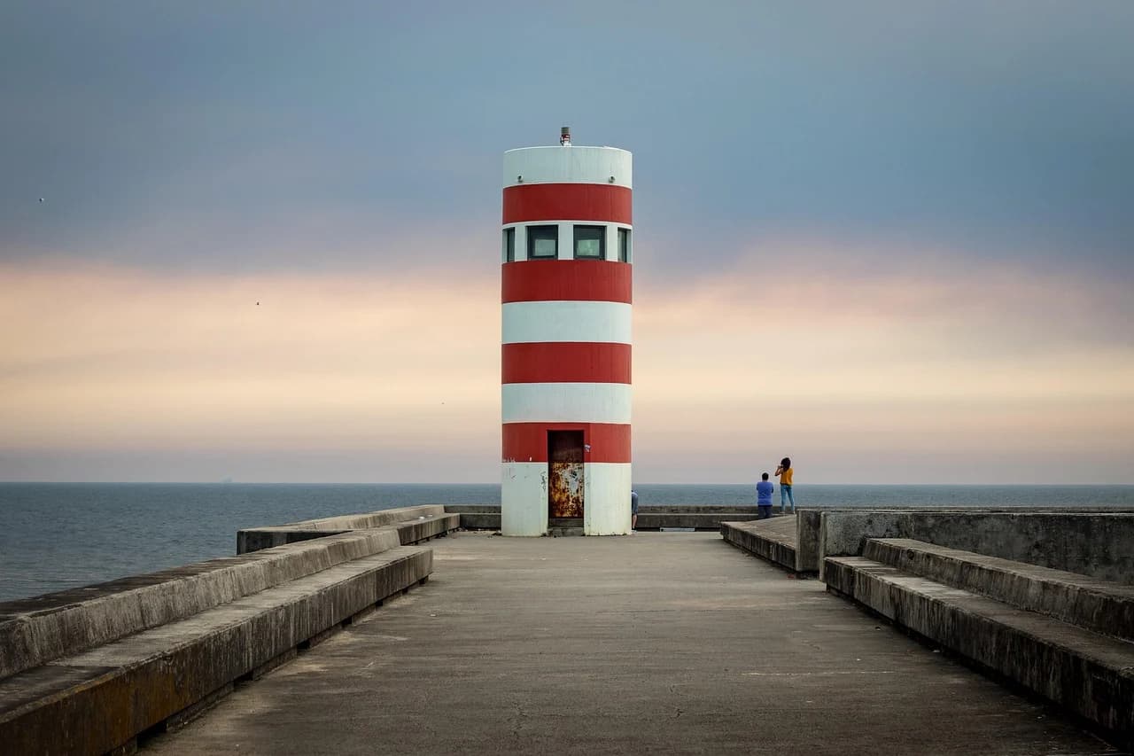 A red and white striped lighthouse stands on a concrete pier, with the sea stretching to the horizon.