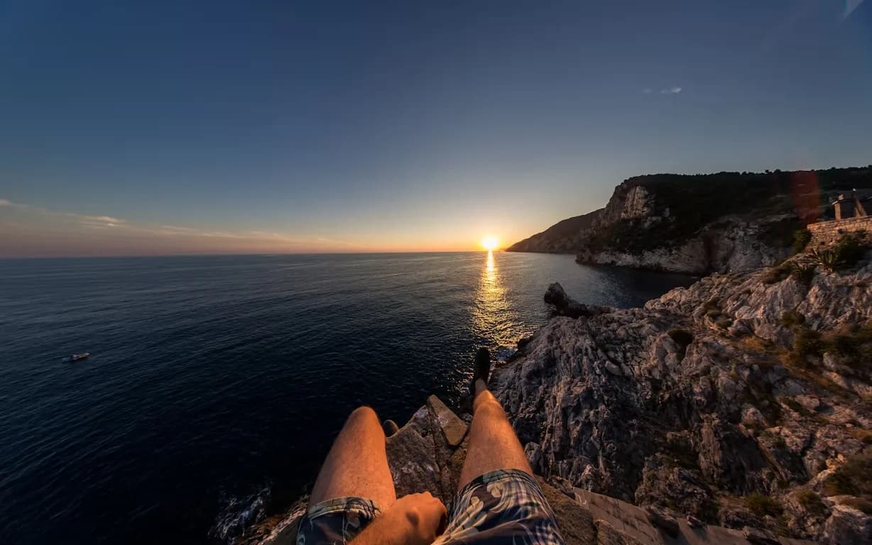 A person sits on the edge of a rocky cliff overlooking the sea, enjoying a beautiful sunset.