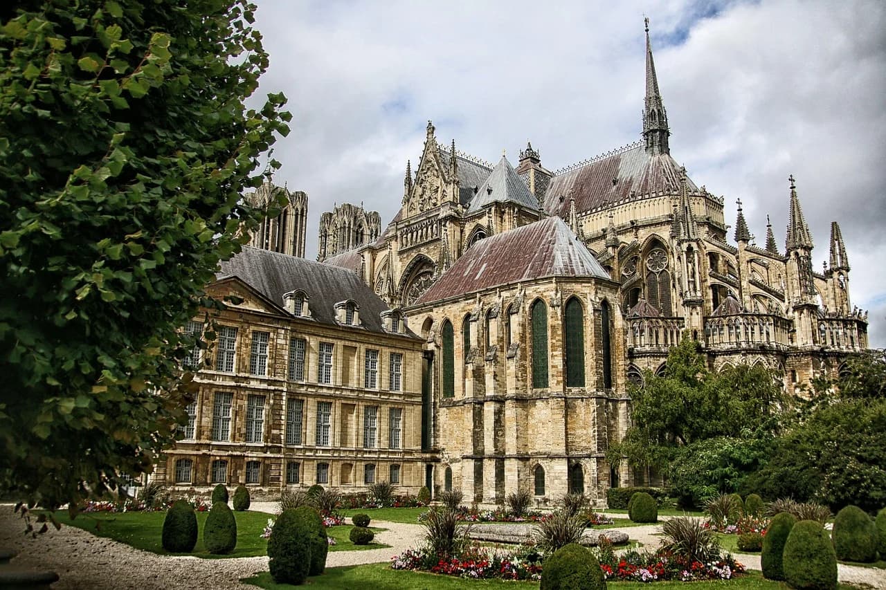 The majestic Reims Cathedral is viewed from a garden with lush green trees, showcasing its intricate stonework and gothic spires.