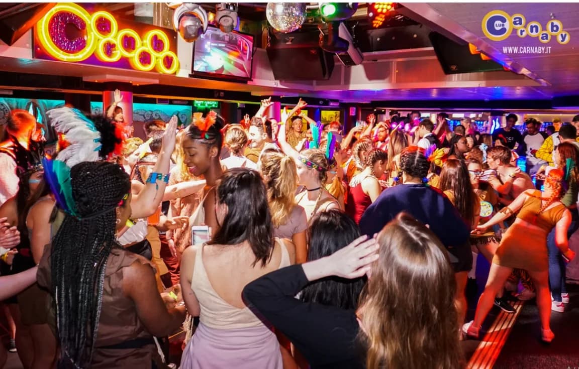 A lively crowd dances under colorful neon lights inside a bustling nightclub.