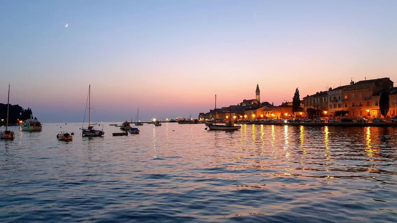 The historic old town of Rovinj is beautifully illuminated at sunset, with its lights and the bell tower of St. Euphemia reflected on the calm sea.