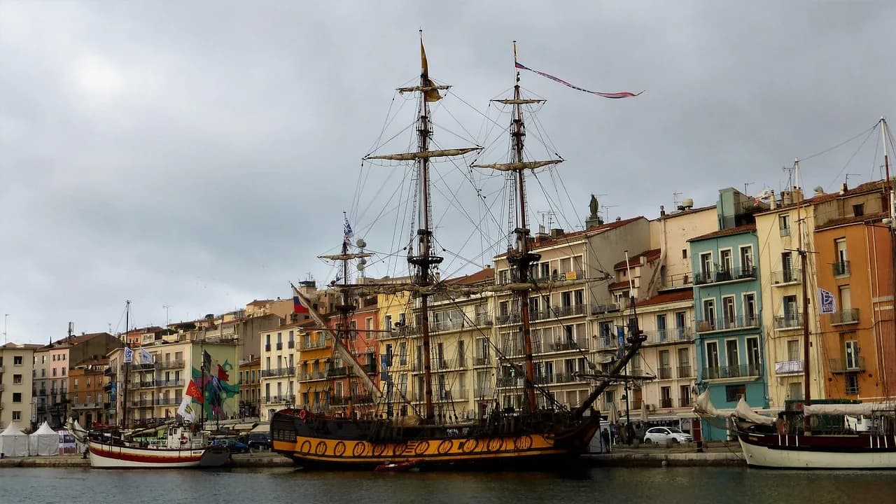 A historic sailing ship is moored in a harbor, with a line of colorful, traditional buildings in the background.