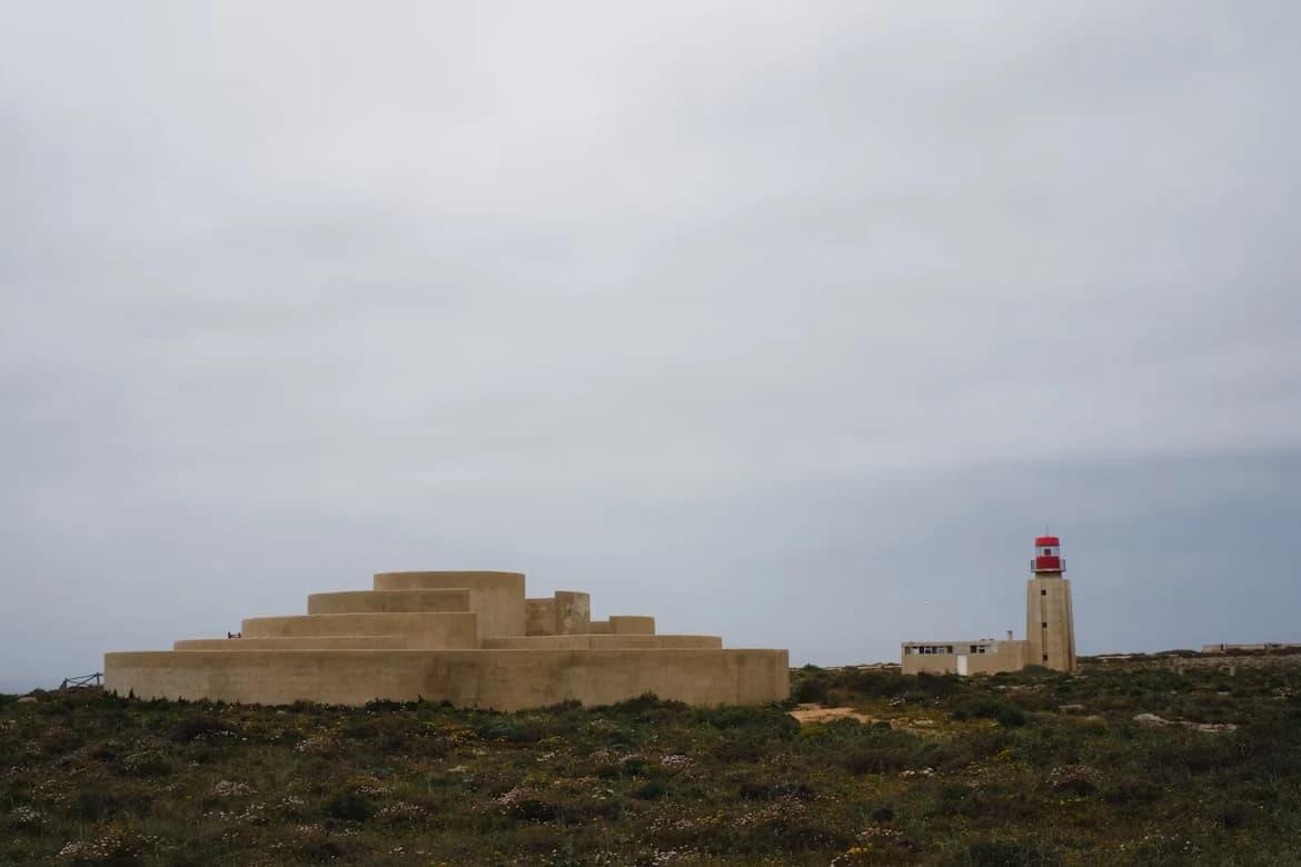 A modern, tiered monument and a lighthouse stand on a rocky, grassy hill, with a cloudy sky in the background.