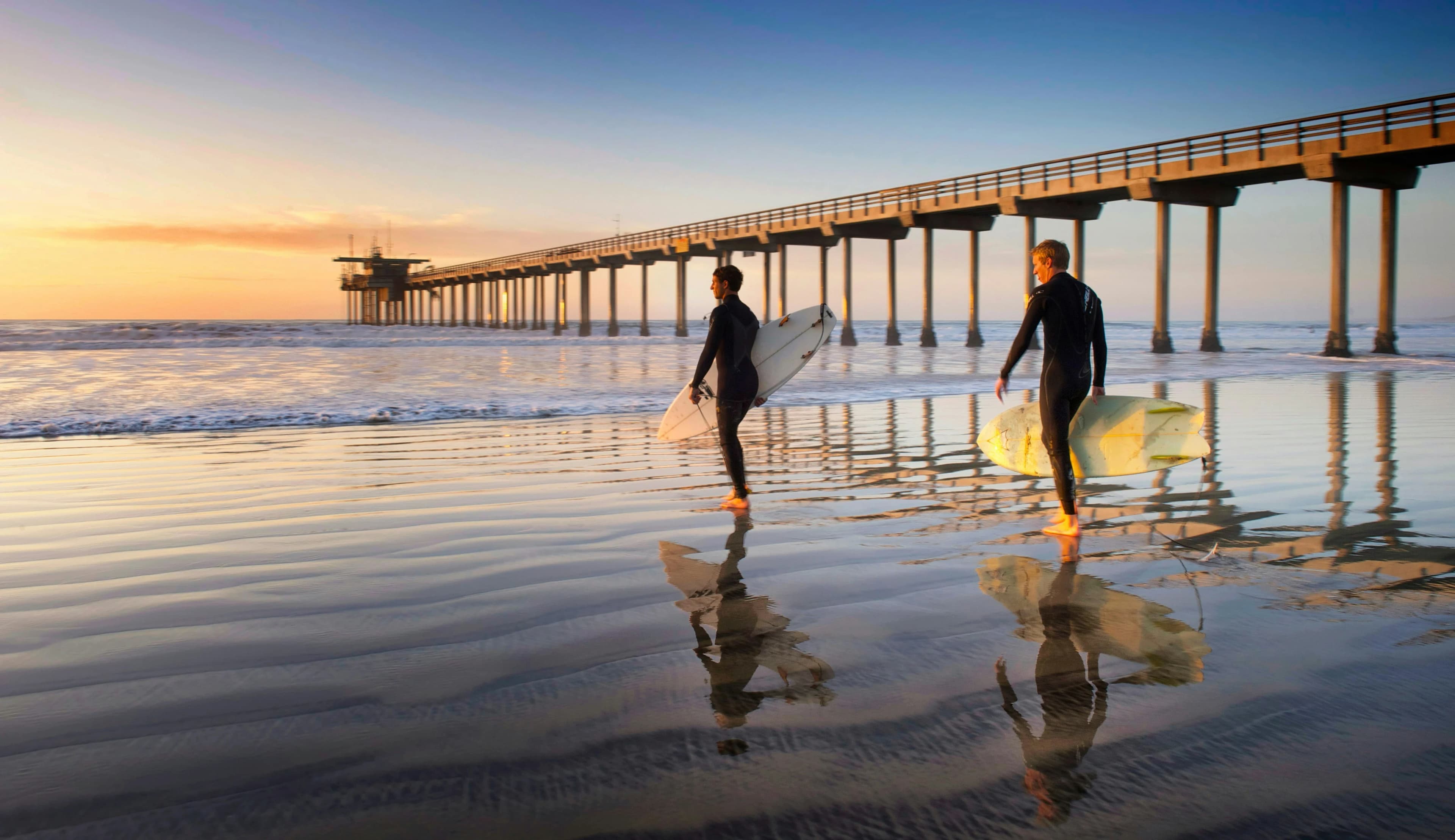 Two surfers walk along the sandy beach at sunset, their silhouettes reflected in the wet sand, with the long Scripps Pier in the background.