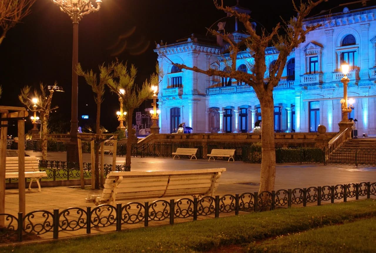 The historic City Hall of San Sebastián is beautifully illuminated in a blue light, with benches and trees in the park in front of it.