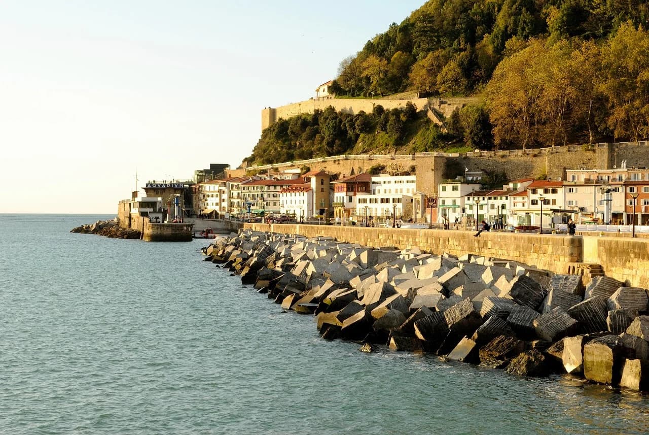The historic buildings of San Sebastián's Old Town are nestled at the base of a green hillside, with a breakwater and the sea in the foreground.