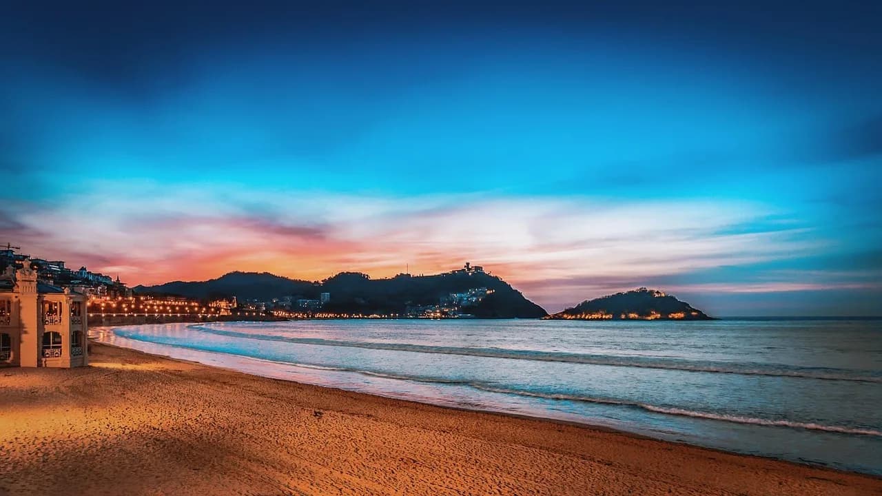 The crescent-shaped La Concha beach is beautifully illuminated at dusk, with the lights of the city and a distant island glowing.