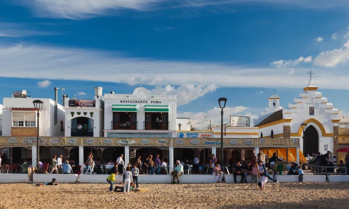 A row of traditional white-washed restaurants and bars lines a sandy beach, with a church steeple and people relaxing in the sun.