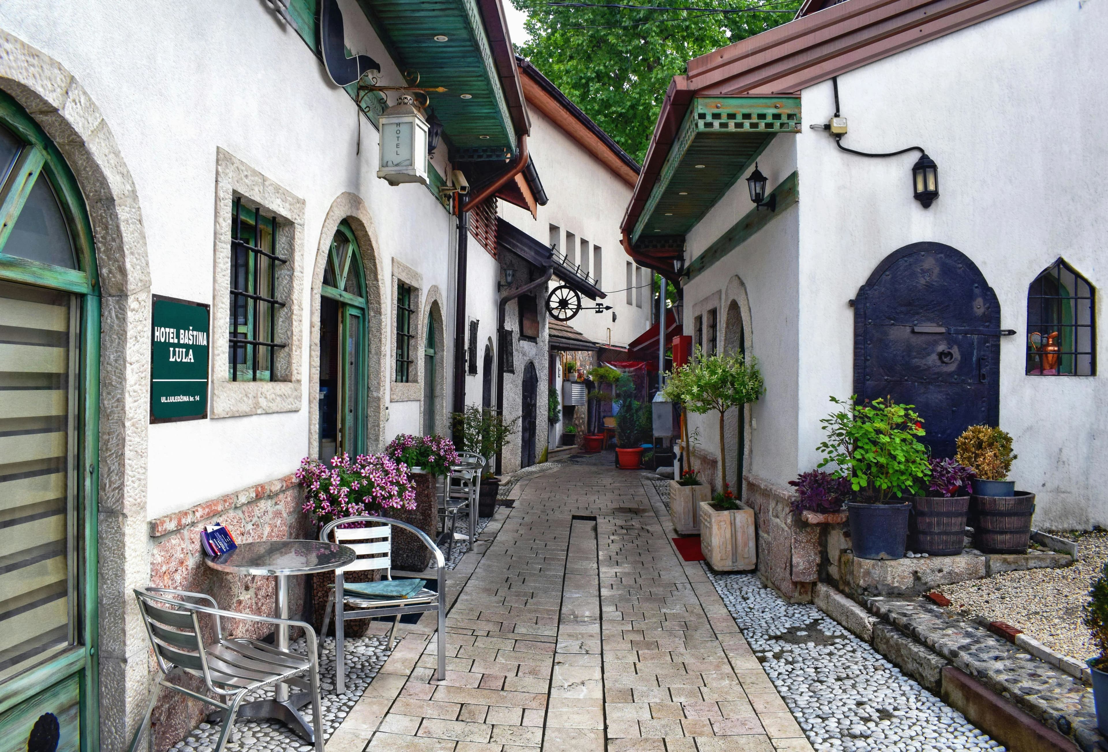 A narrow, cobblestone alleyway in Sarajevo is lined with traditional white-washed buildings and cafes, with lush potted plants and flowers.