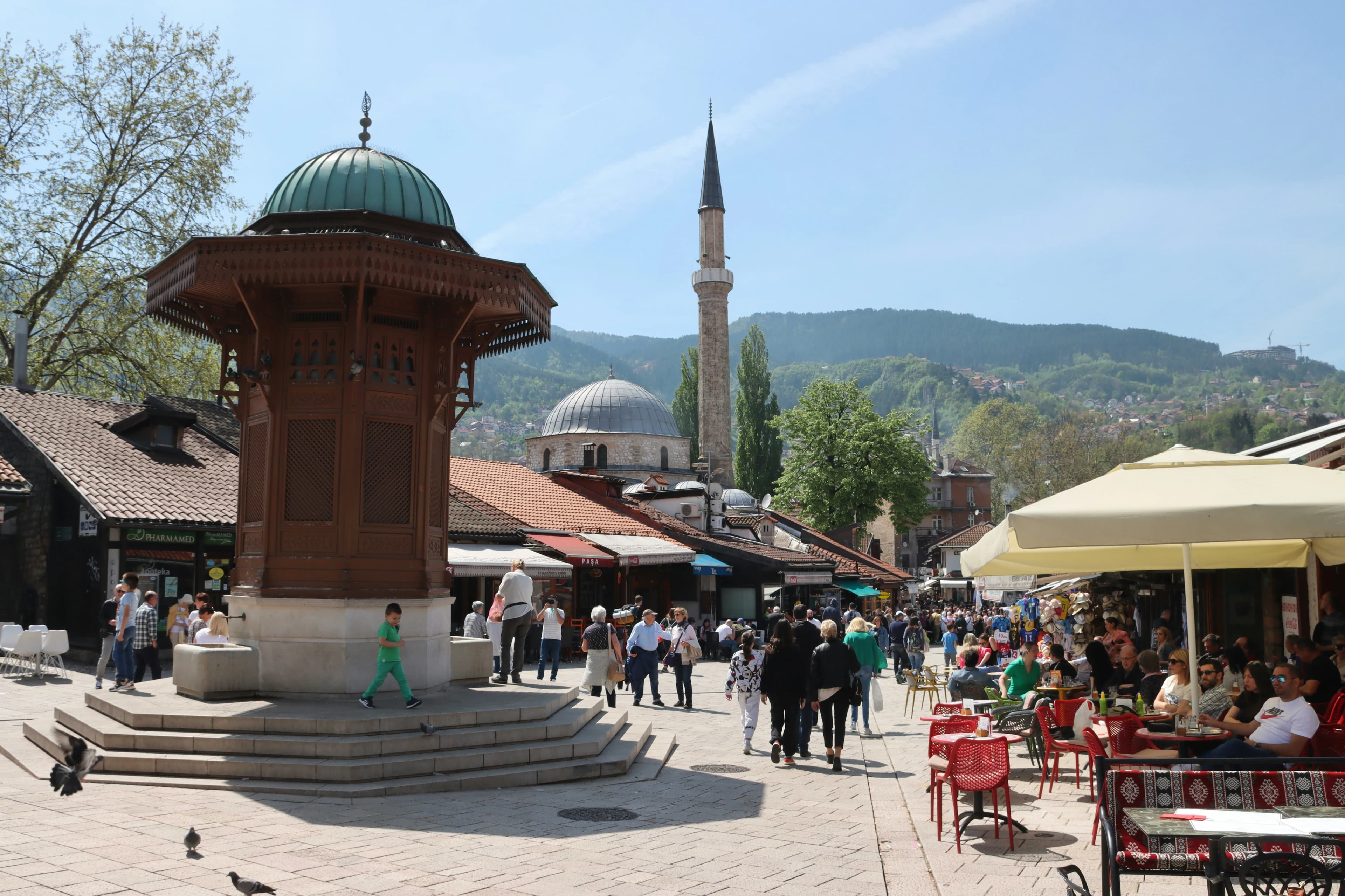 The iconic Sebilj Fountain stands in the center of a bustling city square, with a mosque and a mountain in the background.