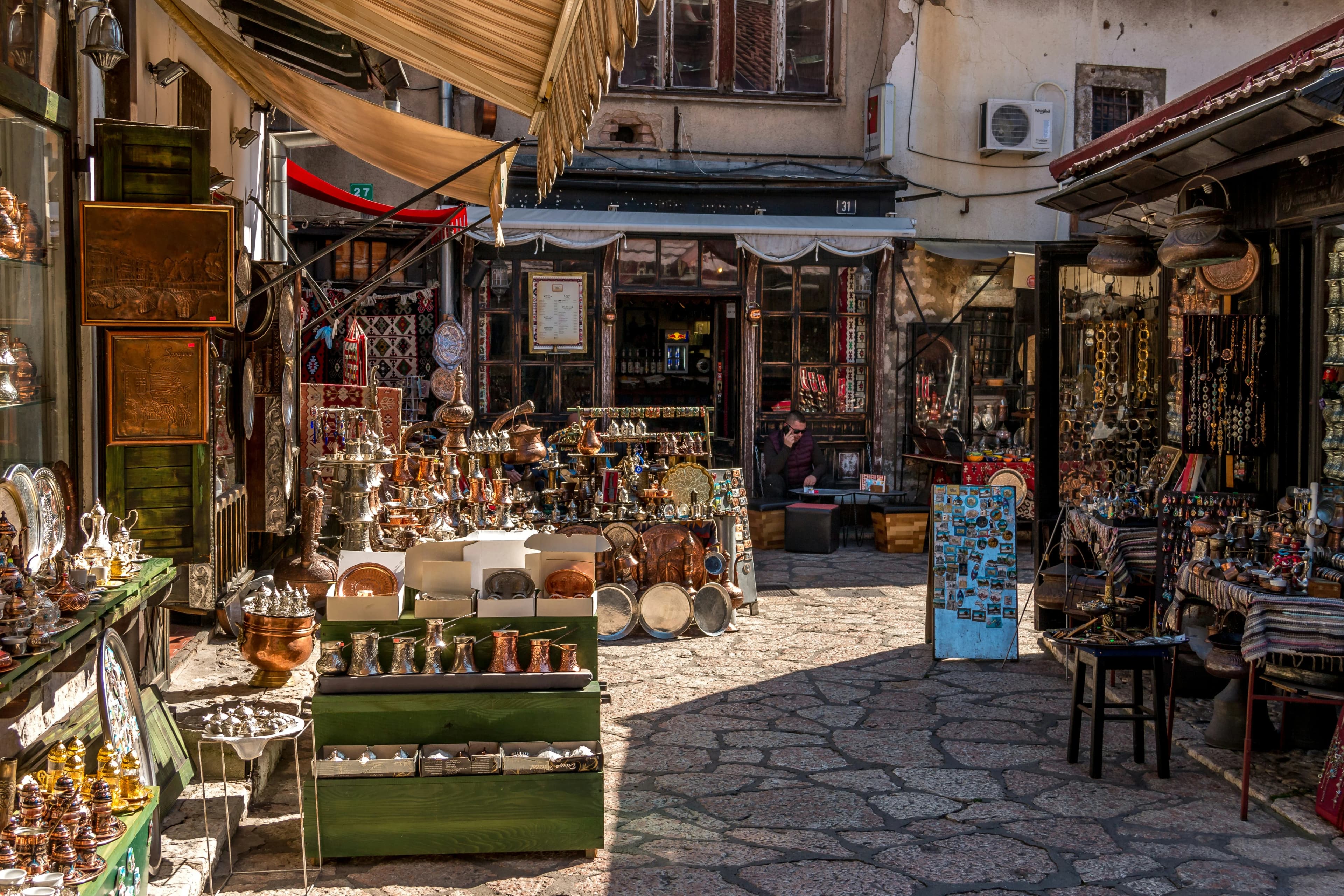 A narrow, bustling market street is filled with stalls selling traditional items and souvenirs, with people walking by and a cafe in the background.