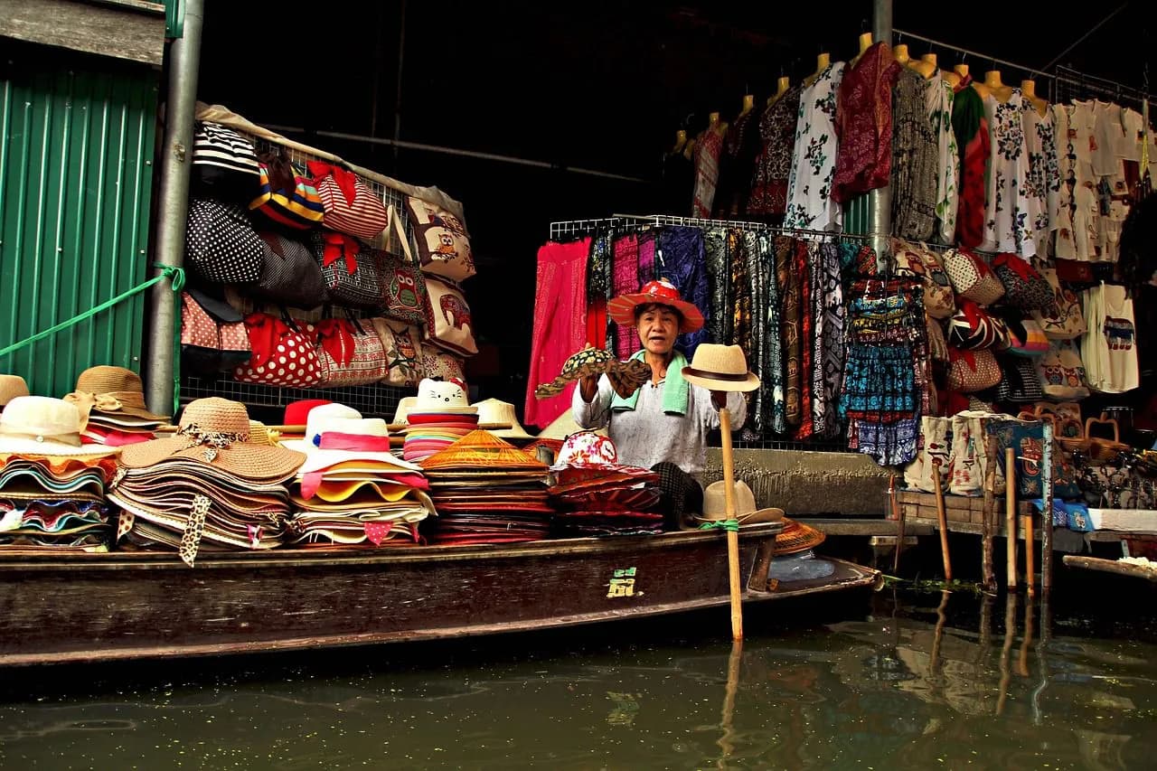A woman in a traditional conical hat sits on a wooden boat filled with colorful hats, bags, and clothes.