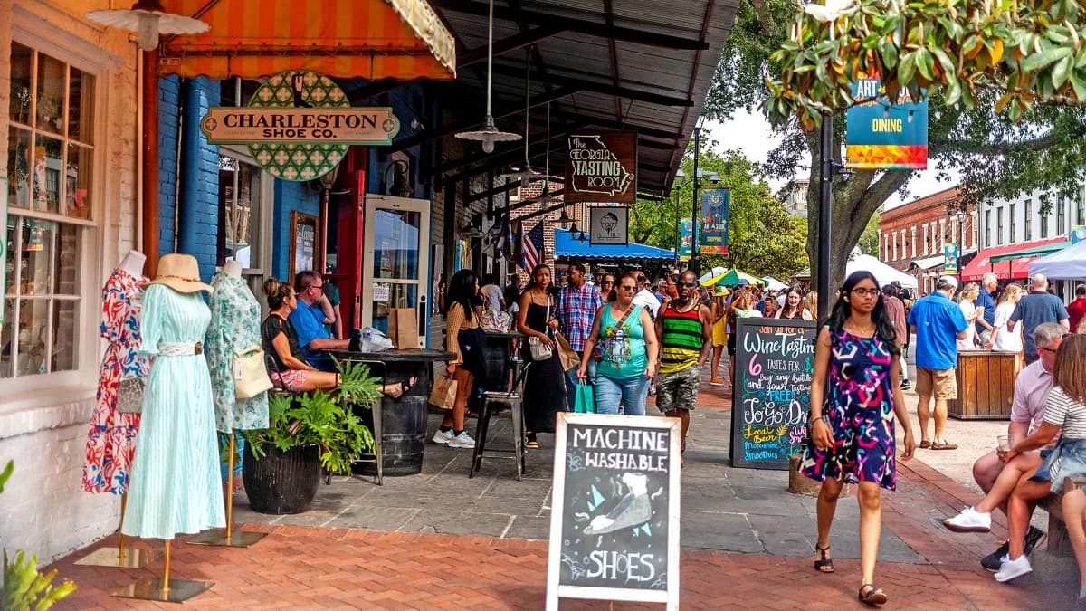 People walk along a historic street lined with shops and restaurants, with a large, traditional building in the background.