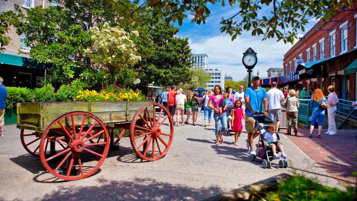 The historic streets of Savannah are bustling with people, with a horse-drawn carriage and colorful storefronts.