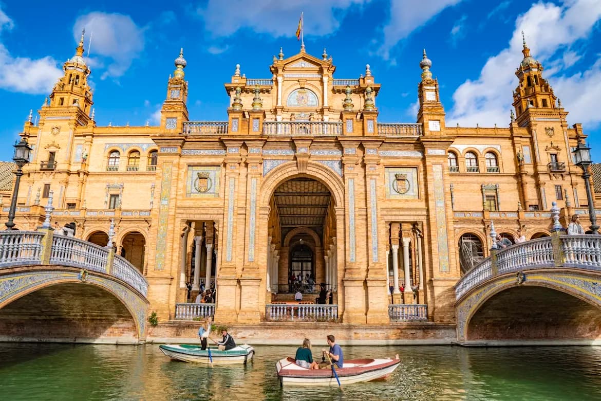 People in small boats row under a bridge in the Plaza de España, with the magnificent building and its numerous arches in the background.