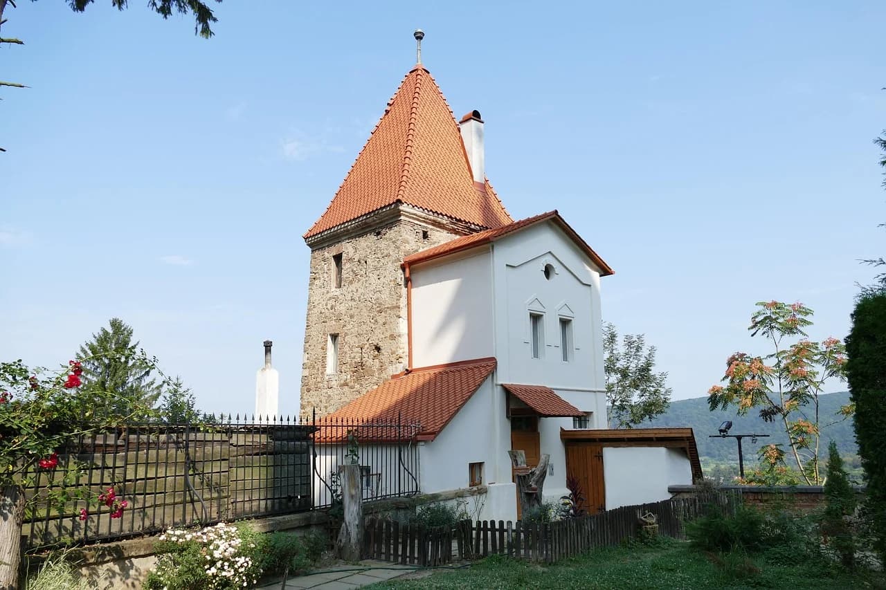 A small, traditional house with a red-tiled roof and a stone tower is nestled in a green garden.