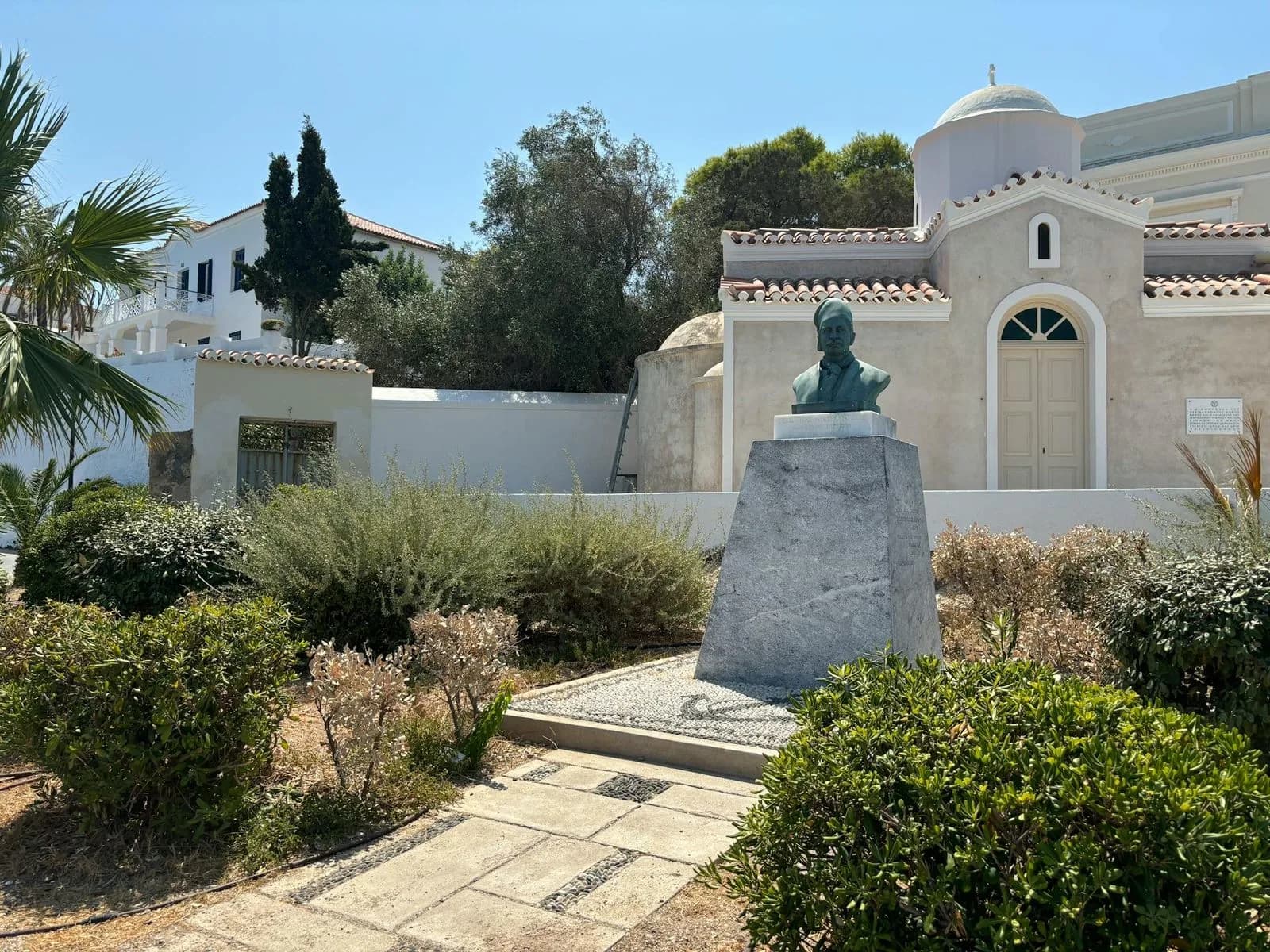 A bronze bust of a historical figure sits on a stone pedestal, with a traditional white chapel in the background.