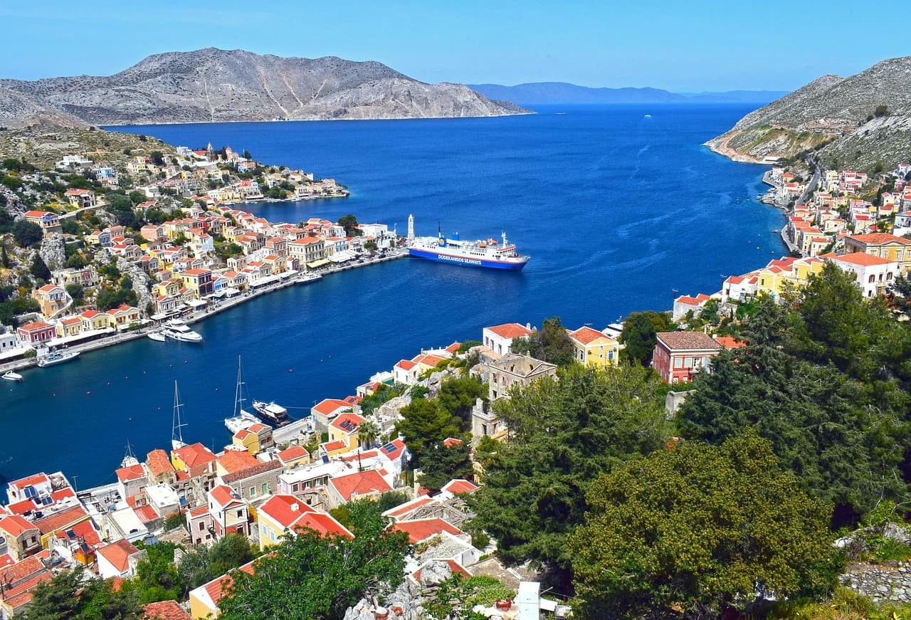 The traditional, colorful buildings of Symi's harbor are reflected on the tranquil water, with fishing boats and yachts moored nearby.