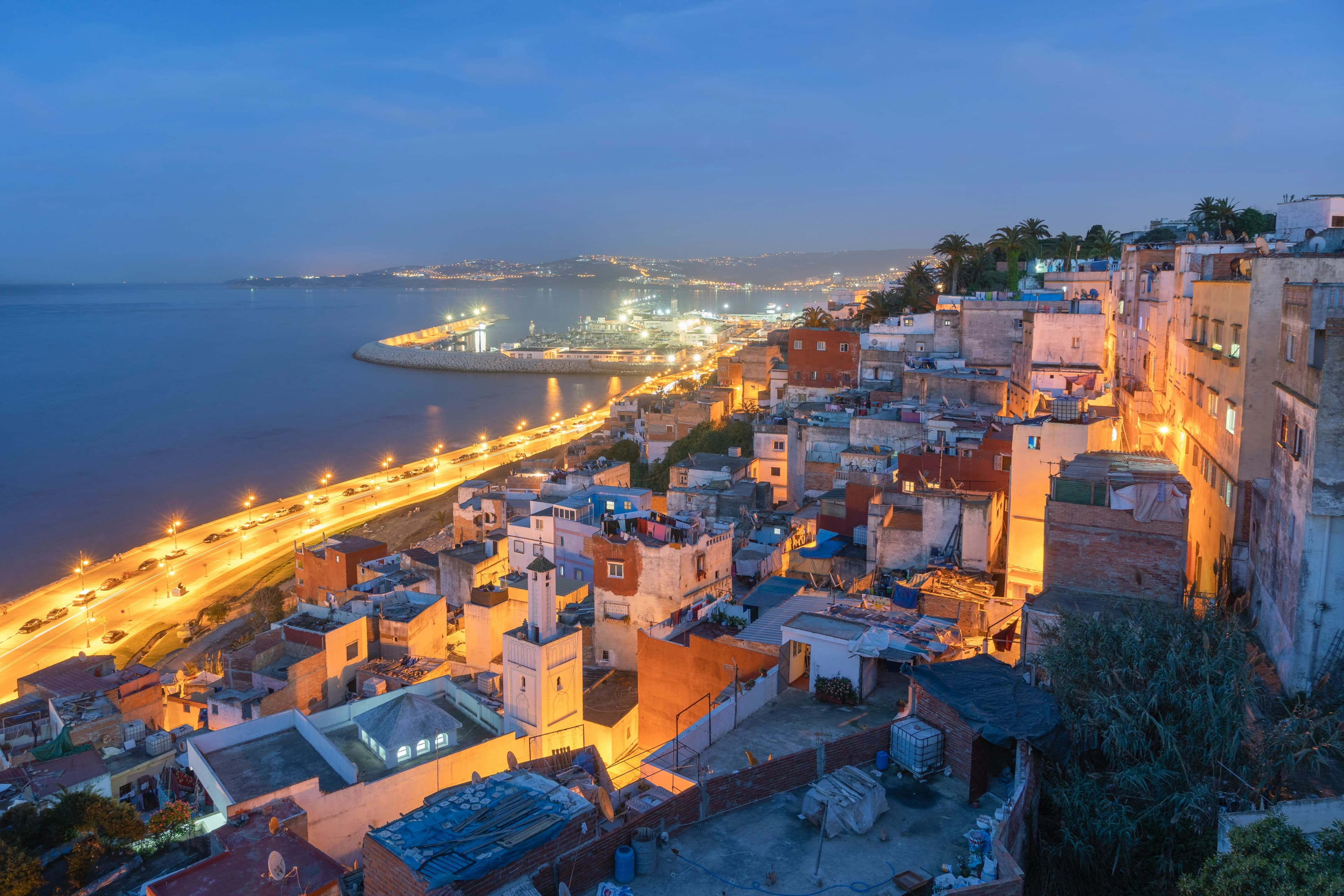 A view of the city of Tangier at dusk, with the lights of the buildings and the port reflecting on the water.