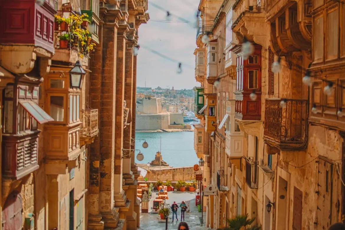 A traditional Maltese street is lined with colorful buildings with unique wooden balconies, with the sea visible at the end of the street.