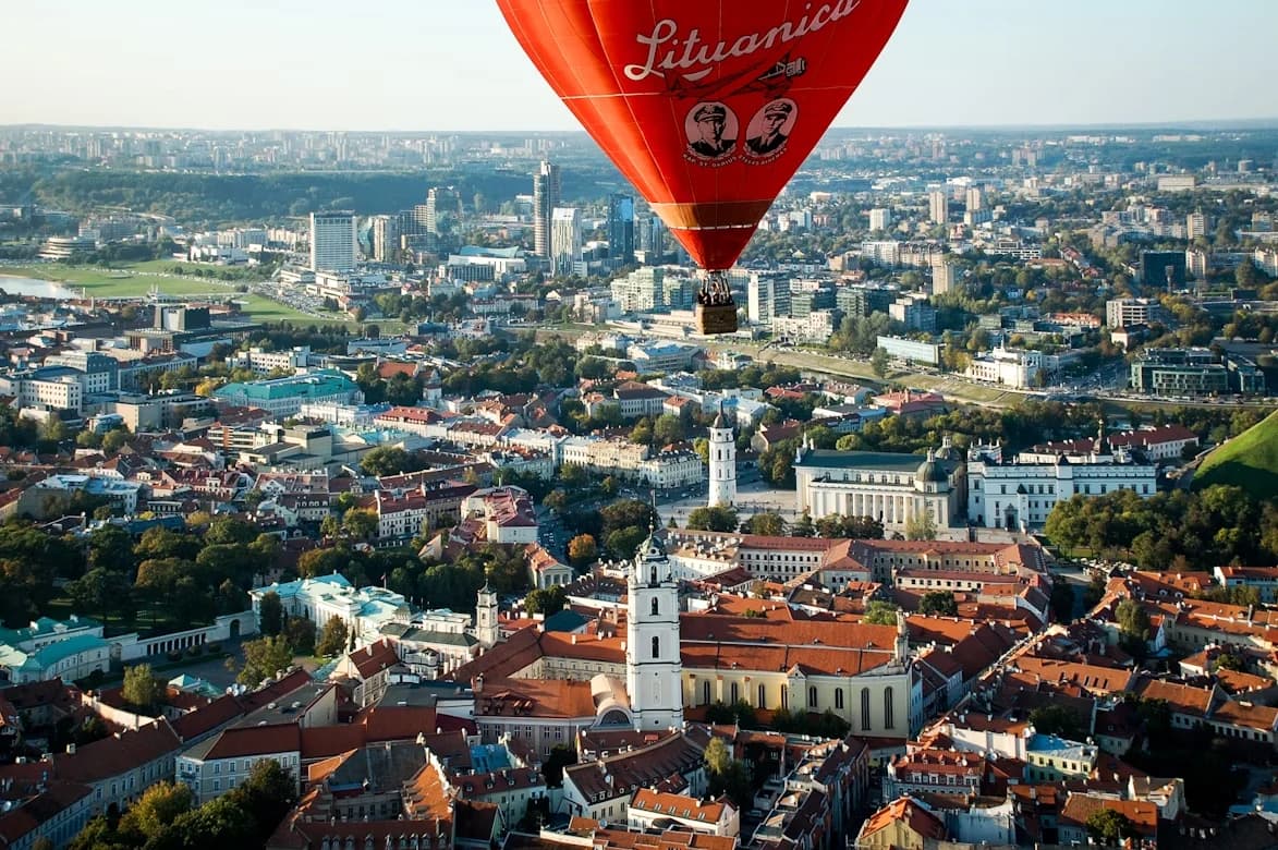 A large, red hot air balloon with "Lituanica" written on it floats over the historic Old Town of Vilnius.