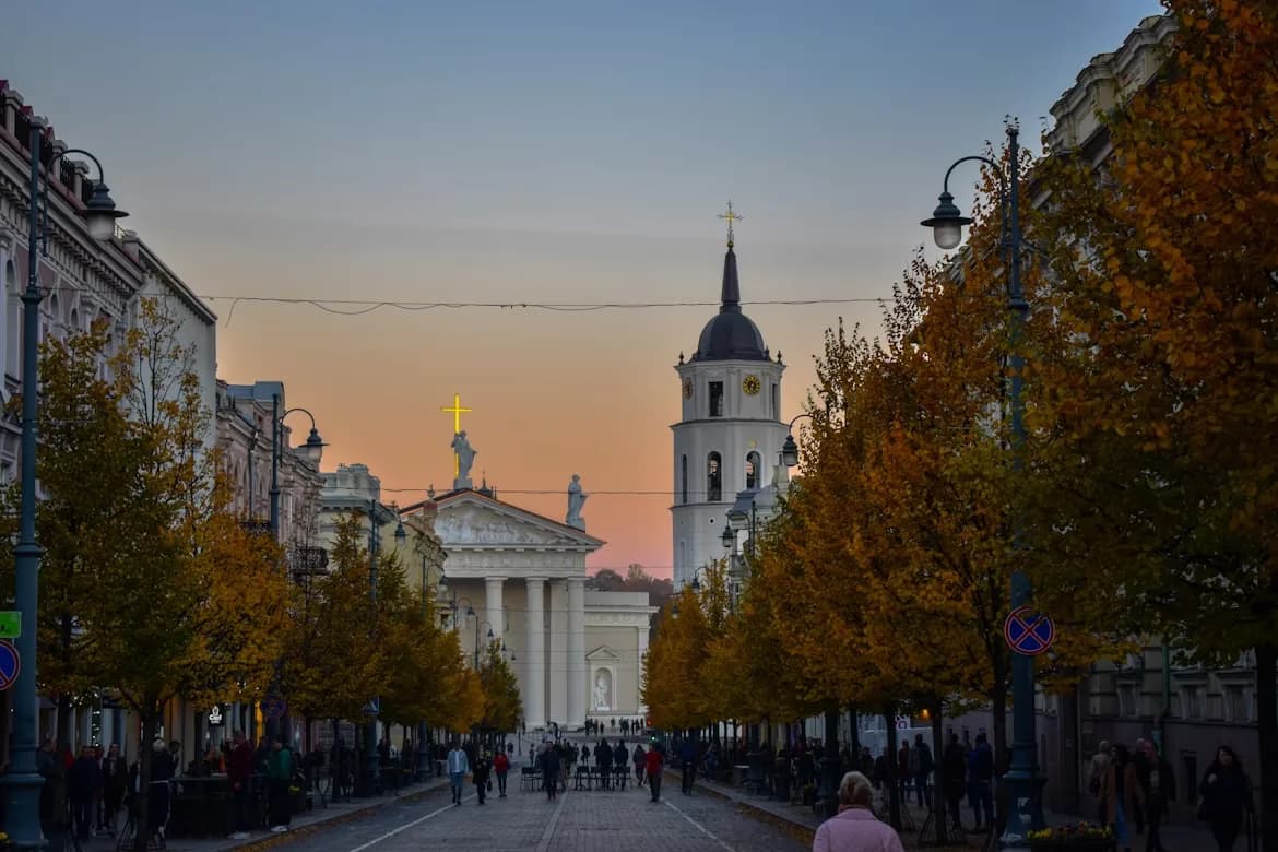The historic Vilnius Cathedral and its bell tower are a beautiful sight on a cobblestone street lined with autumn-colored trees at dusk.